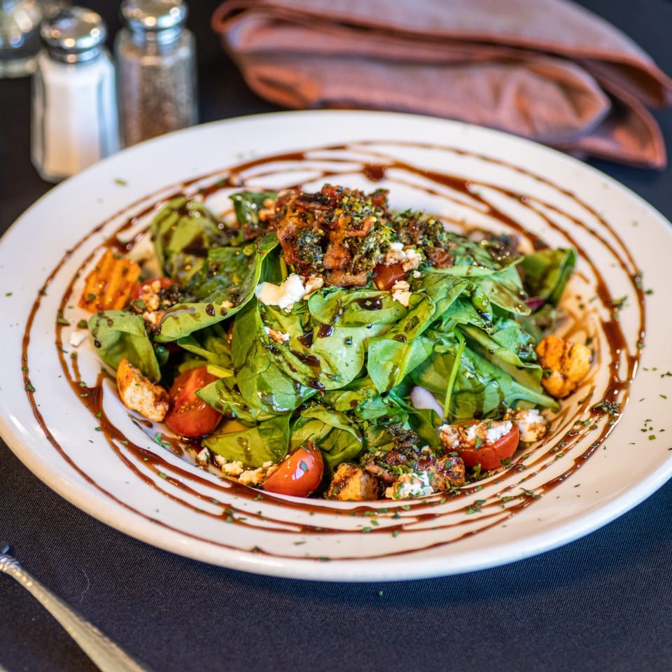 Spinach salad with cherry tomatoes, feta, croutons, and balsamic glaze on a white plate.