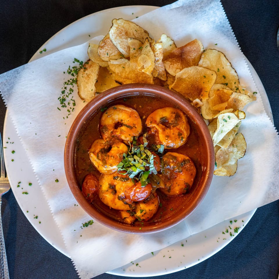 Argentinean shrimp dish in a rustic bowl with red sauce, garnished with herbs, served with potato chips.