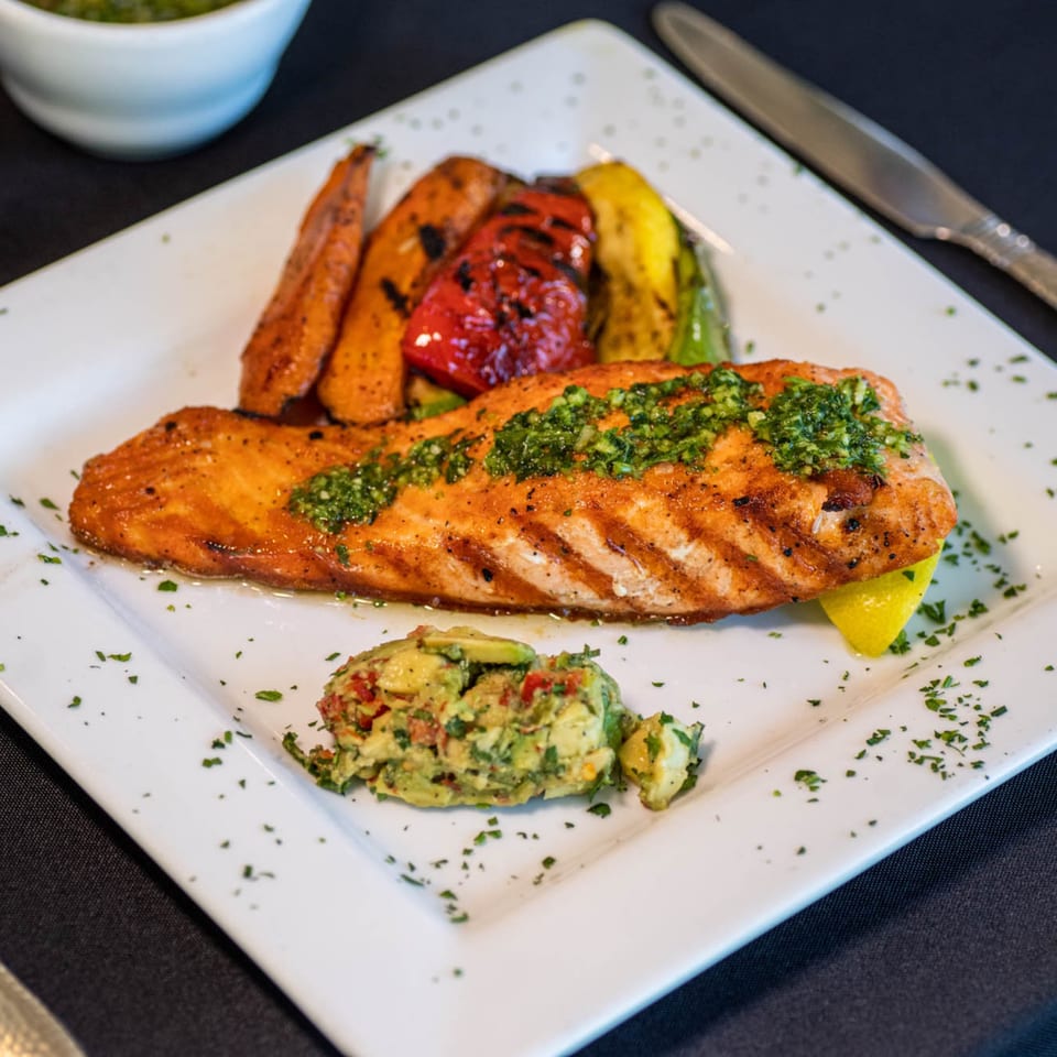 Grilled salmon with chimichurri, roasted vegetables, and avocado salad on a white plate.