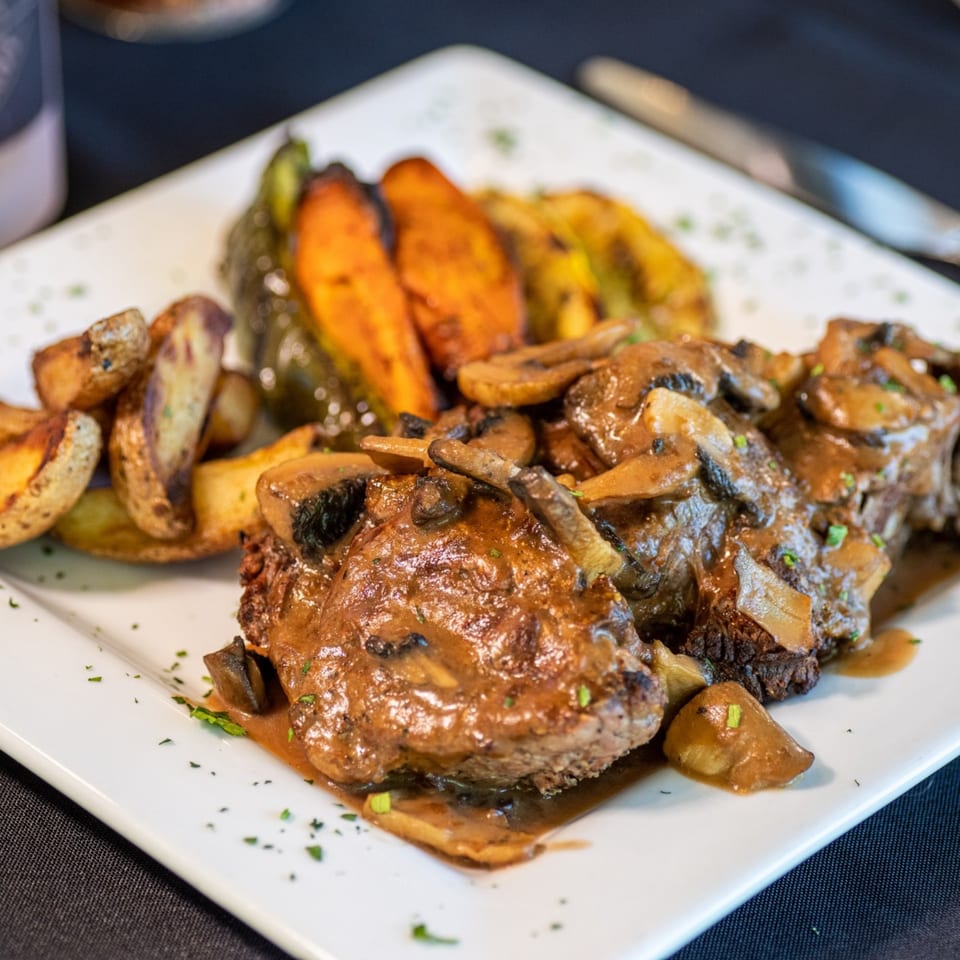 Bendita Cocina's mushroom-smothered steak with roasted potatoes and vegetables on a white plate.