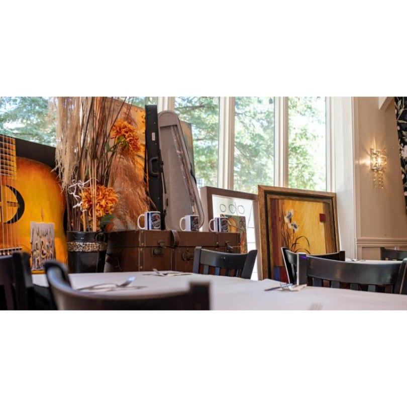 Restaurant interior with tables set, a guitar, artwork, and decorative plants near a large window.
