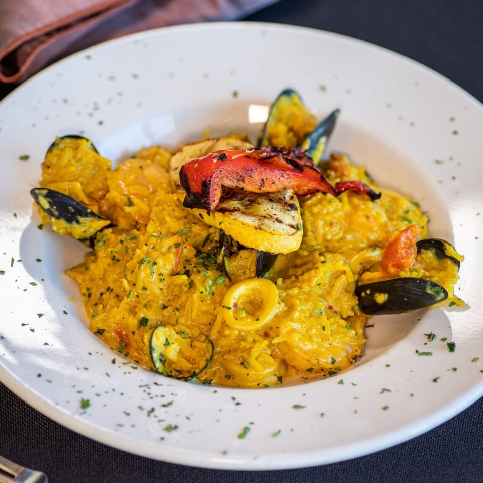 A vibrant yellow seafood paella with mussels, calamari, and grilled vegetables in a white bowl.